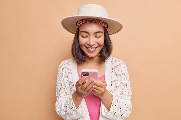 Feliz dama asiática con cabello oscuro usa sombrero y bata blanca mira con alegría la pantalla del teléfono inteligente lee el mensaje de texto contra la pared beige descarga una nueva aplicación