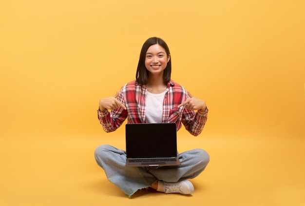 Feliz dama asiática casual señalando con el dedo a una computadora portátil con pantalla negra sentada en el suelo en amarillo