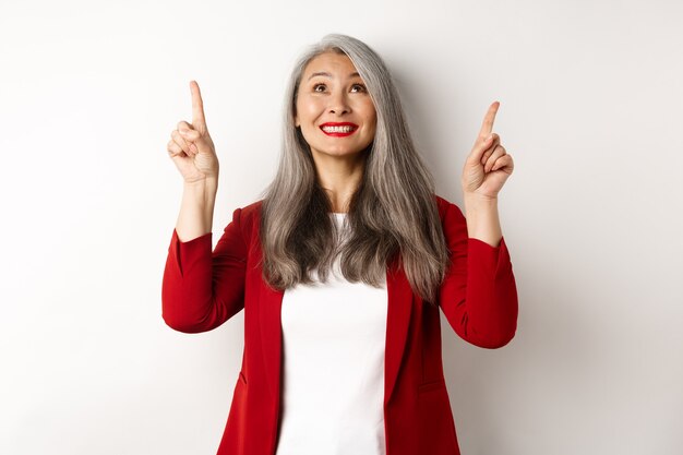 Feliz dama asiática en chaqueta roja y maquillaje, mirando y señalando con el dedo hacia arriba, mostrando una oferta especial, de pie sobre fondo blanco.