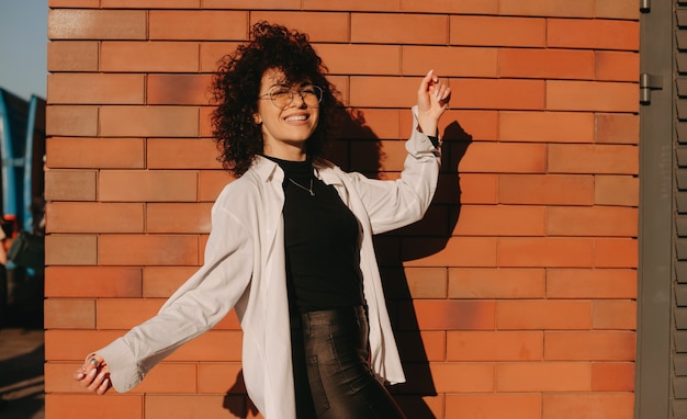 Feliz dama caucásica con el pelo rizado negro lleva anteojos y está posando en un muro de piedra exterior