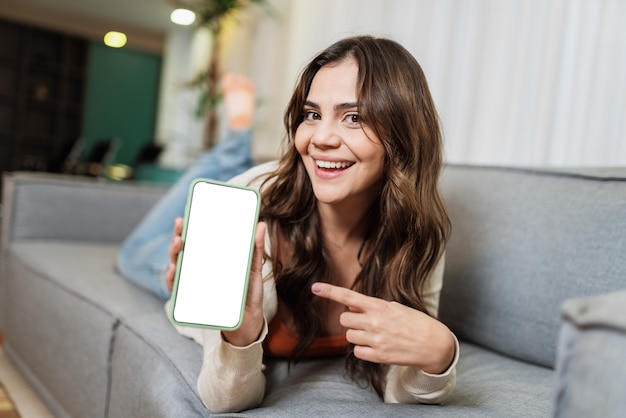 Feliz dama latina relajarse en casa sola en pose de habitación compartir buenas noticias en las redes sociales a través del teléfono celular. Aplicación de teléfono señalando mujer sonriente.