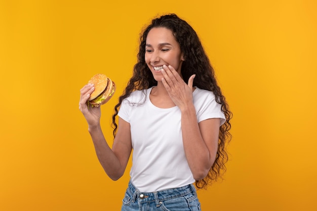 Foto feliz dama latina segurando hambúrguer lambendo o dedo no estúdio