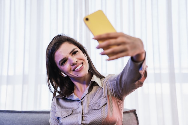 Feliz dama latina sentada en la postura de la habitación y compartir buenas noticias en las redes sociales a través del teléfono celular.