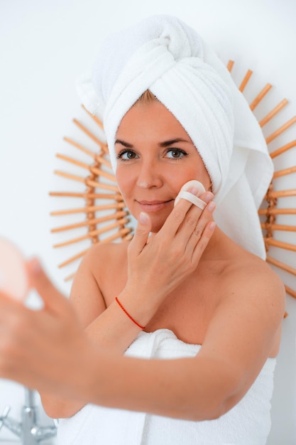 Feliz dama de mediana edad sentada en la encimera del baño empolvándose la cara mientras mira en un espejo pequeño