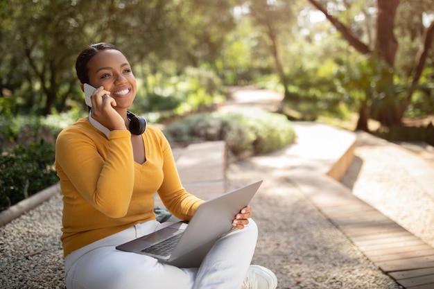 Feliz dama negra relajándose en el parque con una computadora portátil y un estudiante de teléfono celular usando la computadora y hablando