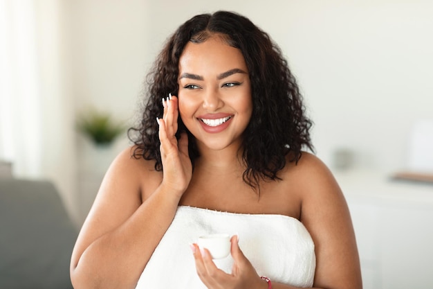 Feliz dama negra con sobrepeso usando crema hidratante para la cara sosteniendo un frasco con productos de belleza y sonriendo