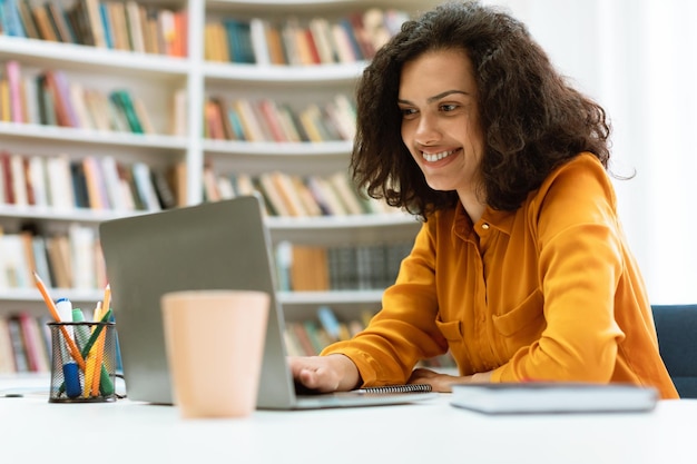 Feliz dama de raza mixta sentada en el escritorio usando una computadora portátil y viendo tutoriales o conferencias estudiando en línea en la biblioteca