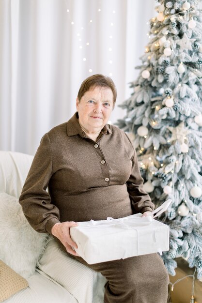 Feliz dama senior en vestido marrón con regalo de Navidad sentado delante de un árbol de Navidad