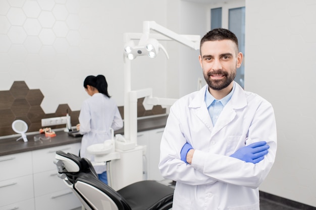 Feliz dentista profesional barbudo joven en guantes y bata blanca de pie delante de la cámara en el fondo de su asistente