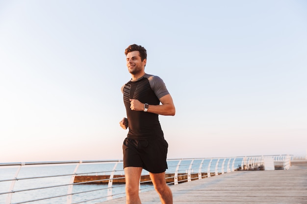 Feliz desportista de Bermuda e camiseta correndo ao longo do cais ou calçadão à beira-mar, durante o nascer do sol