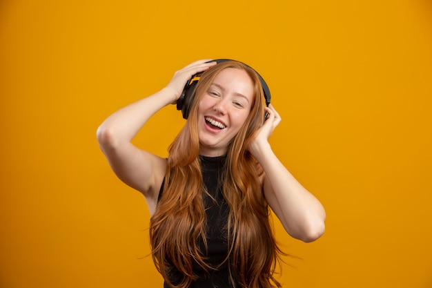 Feliz y despreocupada, emocionada chica pelirroja inconformista como sus nuevos auriculares, bailando con las manos levantadas y sonriendo disfruta escuchando música sobre la pared naranja, aumenta el estado de ánimo con la canción favorita.