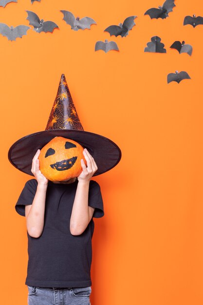 Feliz Dia das Bruxas. Crianças assustadoras de halloween. Menino com um chapéu segurando uma abóbora de halloween na frente de sua cabeça em um fundo laranja com morcegos de papel, moldura vertical. Travessuras ou travessuras conceito de halloween.