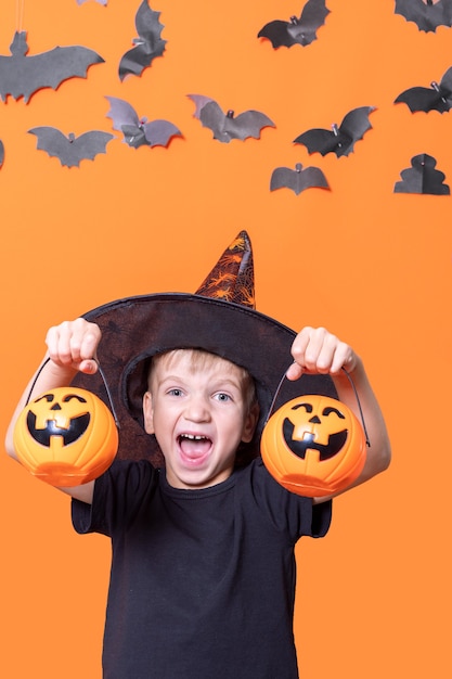 Feliz Dia das Bruxas. Crianças assustadoras de halloween. Menino com um chapéu segurando uma abóbora de halloween na frente de sua cabeça em um fundo laranja com morcegos de papel, moldura vertical. Travessuras ou travessuras conceito de halloween.