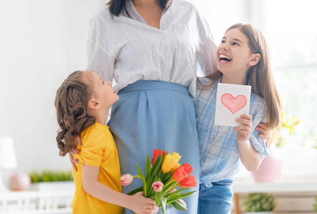 Feliz Dia das Mães! As filhas das crianças estão parabenizando a mãe e dando-lhe cartão postal e flores. Mamãe e meninas sorrindo e abraçando. Férias em família e união.