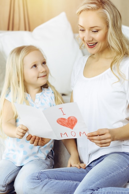 Feliz dia das mães em apartamento ensolarado! Filha criança felicita a mãe e dá seu cartão postal com desenho de coração. Conceito de família.