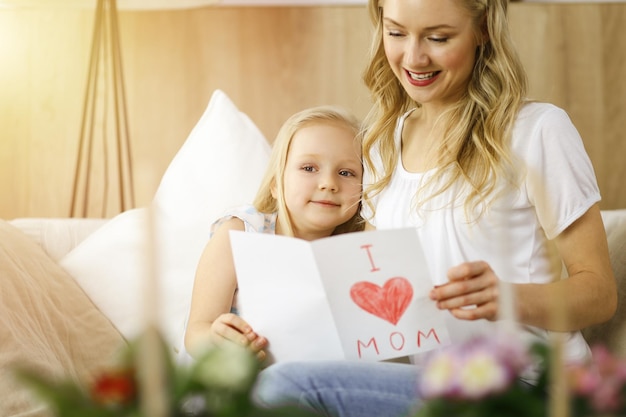Feliz dia das mães em apartamento ensolarado. Filha criança felicita a mãe e dá seu cartão postal com desenho de coração. Conceito de família.