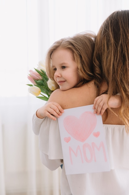 Feliz Dia das Mães. Filha criança felicita mães e dá-lhe um cartão postal e flores tulipas.