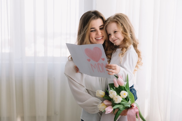 Feliz Dia das Mães. Filha criança felicita mães e dá-lhe um cartão postal e flores tulipas.