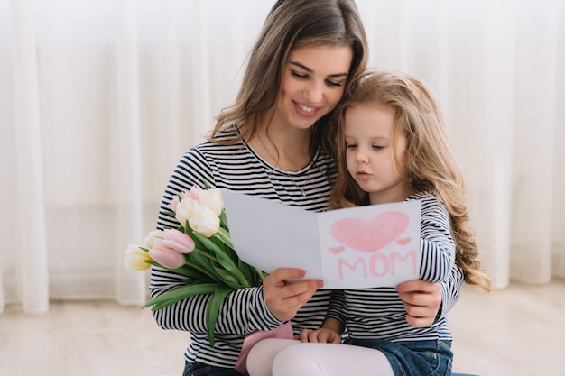 Feliz Dia das Mães. Filha criança felicita mães e dá-lhe um cartão postal e flores tulipas.