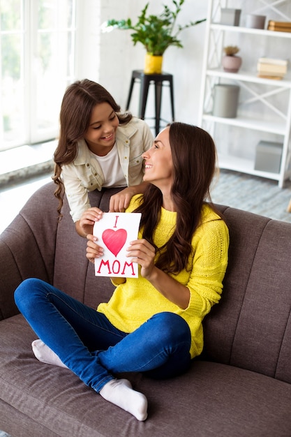 Feliz Dia das Mães. Filha fofa parabeniza a mãe dela e dá a ela um cartão postal. Mãe e menina sorrindo e se abraçando no sofá. Férias em família e união