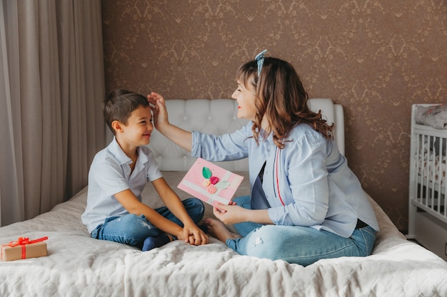 Feliz dia das mães. filho parabeniza a mãe e dá um cartão postal. mãe e bebê sorriem e se abraçam. feriado familiar.