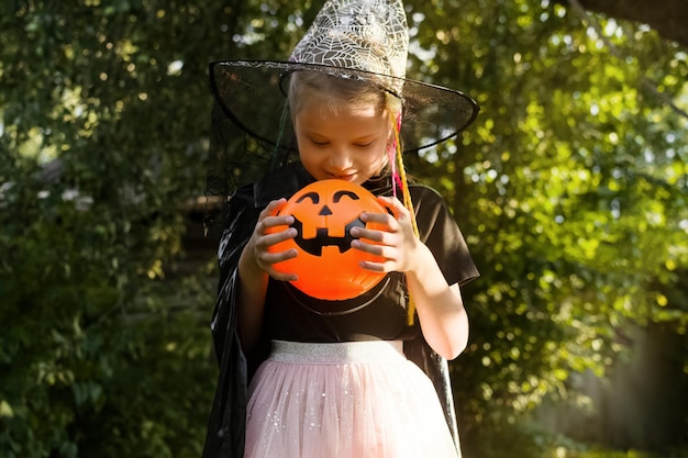 Feliz dia do dia das bruxas menina vestida com uma fantasia de bruxa segura um balde de doces e olha para doces