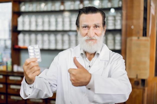 Feliz dia do farmacêutico Sorrindo alegre farmacêutico sênior masculino em casaco branco em pé na antiga farmácia vintage segurando blister com comprimidos e mostrando o polegar para cima