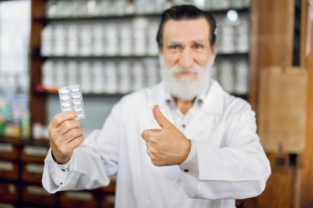 Feliz dia do farmacêutico Visão desfocada do sorridente farmacêutico sênior alegre em jaleco branco em pé na antiga farmácia vintage segurando blister com comprimidos e mostrando o polegar para cima