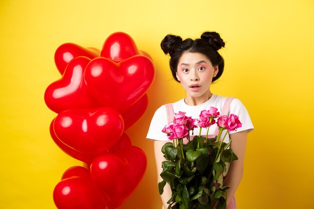 Feliz dia dos namorados. menina asiática surpresa recebe buquê de rosas cor de rosa do amante, olhando com admiração para a câmera, em pé perto de balões de coração vermelho, fundo amarelo.