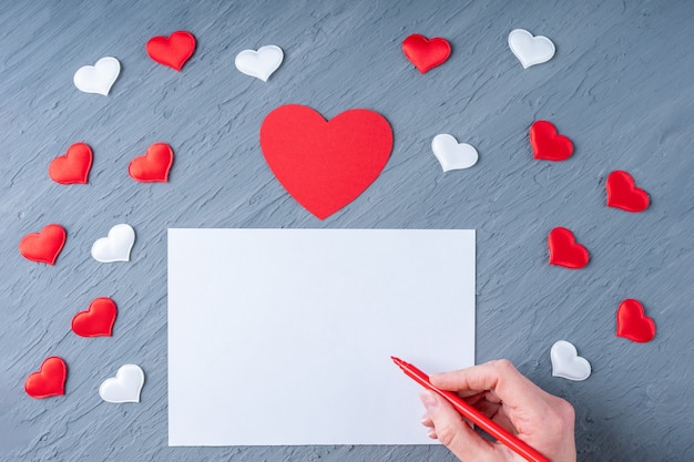 Feliz dia dos namorados saudações. Mão segura uma caneta vermelha para escrever uma carta de amor ou parabéns sobre um fundo cinza