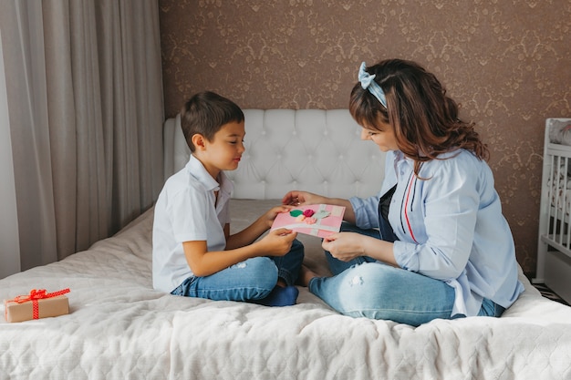 Feliz día de la madre. Hijo felicita a mamá y le da una postal. Mamá y bebé sonríen y se abrazan. Vacaciones familiares.