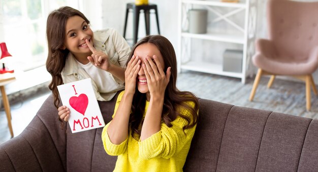 Feliz día de la madre. Linda hija felicita a su mamá y le da una postal. Mamá y niña sonriendo y abrazándose en el sofá. Vacaciones familiares y convivencia