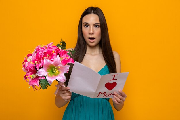 Feliz día de la madre. Mujer joven hermosa sorprendida que sostiene la tarjeta de felicitación y el ramo