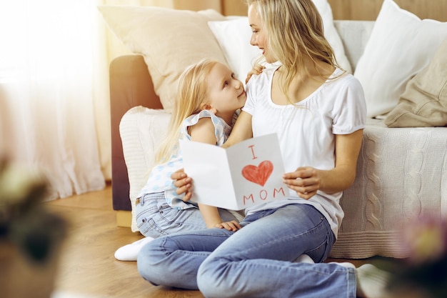 ¡Feliz día de la madre en piso soleado! La hija del niño felicita a mamá y le da una postal con un dibujo del corazón. concepto de familia.