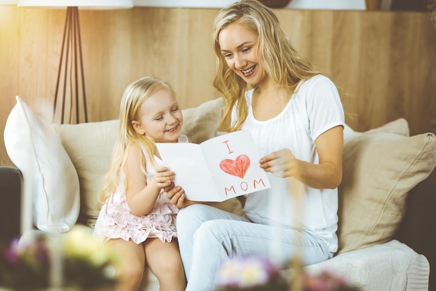 Feliz día de la madre en piso soleado. La hija del niño felicita a mamá y le da una postal con un dibujo del corazón. concepto de familia