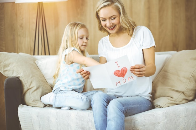 Feliz día de la madre en piso soleado. La hija pequeña felicita a mamá y le da una postal con un dibujo de corazón. Concepto de familia