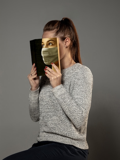 Feliz día mundial del libro 2020, esté seguro y lea para convertirse en otra persona: una mujer que cubre la cara con un libro en una máscara mientras lee sobre un fondo gris de estudio. Celebración, educación, arte, concepto de protección.