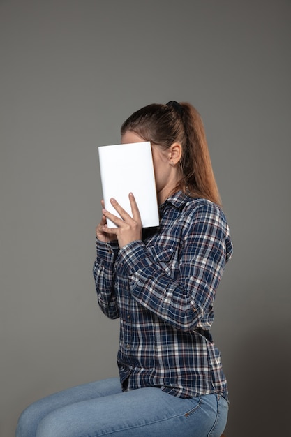 Feliz día mundial del libro y los derechos de autor, lea para convertirse en otra persona: una mujer que cubre la cara con un libro mientras lee en una pared gris.