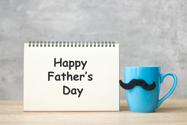 Feliz día del padre con bloc de notas de papel Taza de café azul o taza de té y decoración de bigote negro en la mesa Día internacional del hombre y concepto de celebración