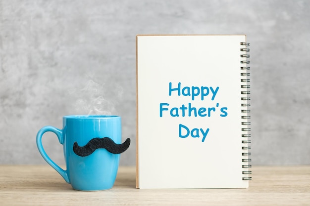 Feliz día del padre con bloc de notas de papel Taza de café azul o taza de té y decoración de bigote negro en la mesa Día internacional del hombre y concepto de celebración