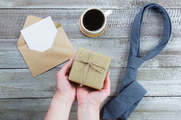 Foto feliz día del padre. manos de mujer con caja de regalo o presente. corbata azul, taza de café y vacía en blanco.