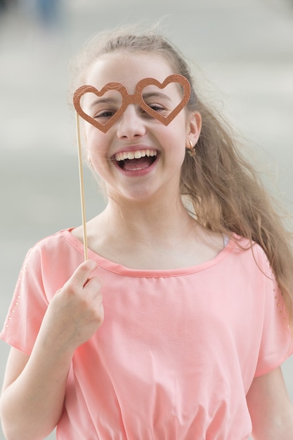 Feliz día de San Valentín. Niño pequeño con sonrisa feliz y mirada divertida a través de gafas en forma de corazón. Niña feliz sonriendo con accesorios de fotomatón en palo. Celebrando el feliz día internacional del niño.