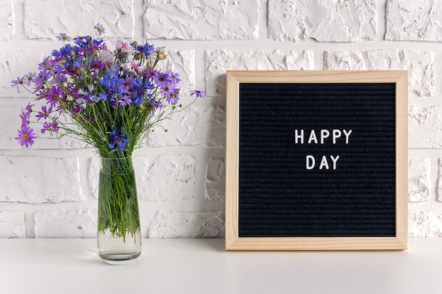 Feliz día texto en pizarra negra y ramo de flores azules en florero sobre la mesa contra la pared de ladrillo blanco