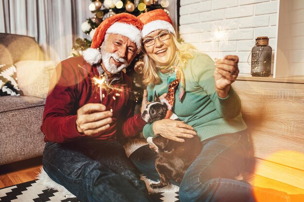 Feliz y divertida pareja senior sosteniendo bengalas y celebrando el año nuevo junto con su adorable perro Boston terrier.