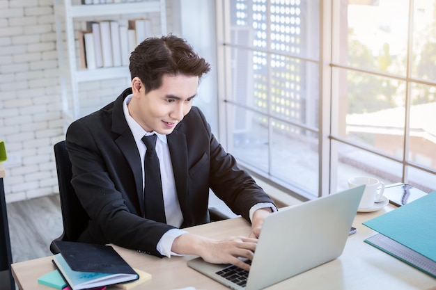 Feliz do jovem empresário asiático ver um plano de negócios de sucesso no computador laptop e uma caneta no fundo da mesa de madeira no escritório, o negócio expressa confiança encoraja