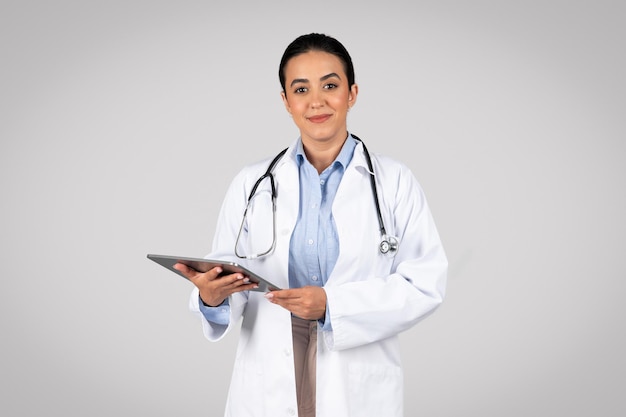 Feliz doctora brasileña con bata blanca tomando notas en una tableta usando una aplicación para chatear posando sobre gris