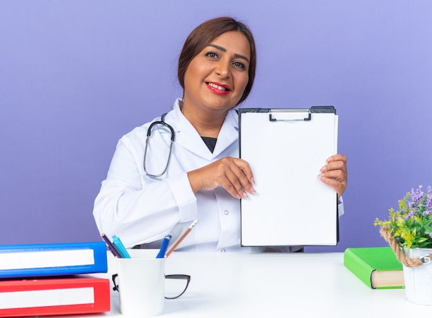 Feliz doctora de mediana edad en bata blanca con estetoscopio mostrando portapapeles con páginas en blanco mirando al frente sonriendo alegremente sentado en la mesa sobre pared azul