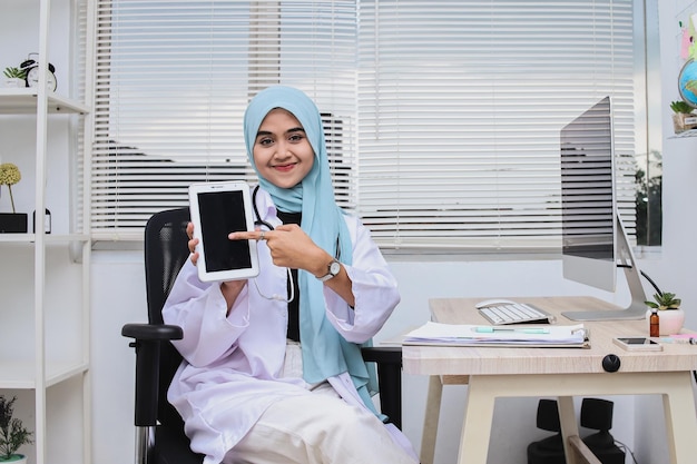 Feliz doctora mujer musulmana en hijab mostrando tableta digital con pantalla en blanco, maqueta, copia spa