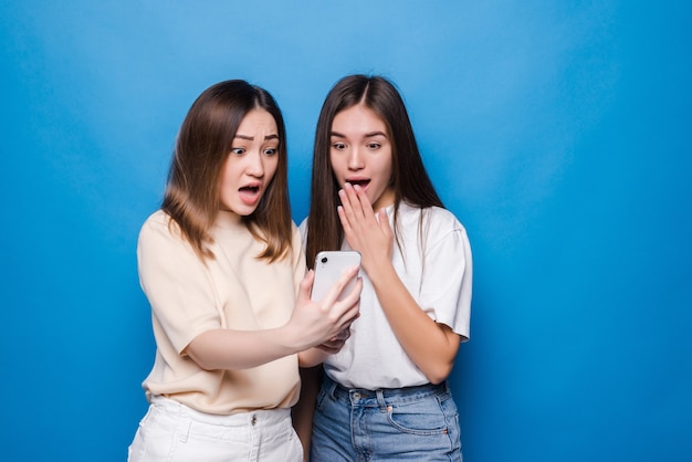 Feliz dos niñas riendo y señalando con el dedo a la pantalla del teléfono inteligente mientras toman selfie aislado sobre pared azul