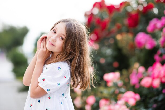 Feliz e alegre garotinha adorável parada no jardim do parque sobre arbustos florescentes de flor rosa